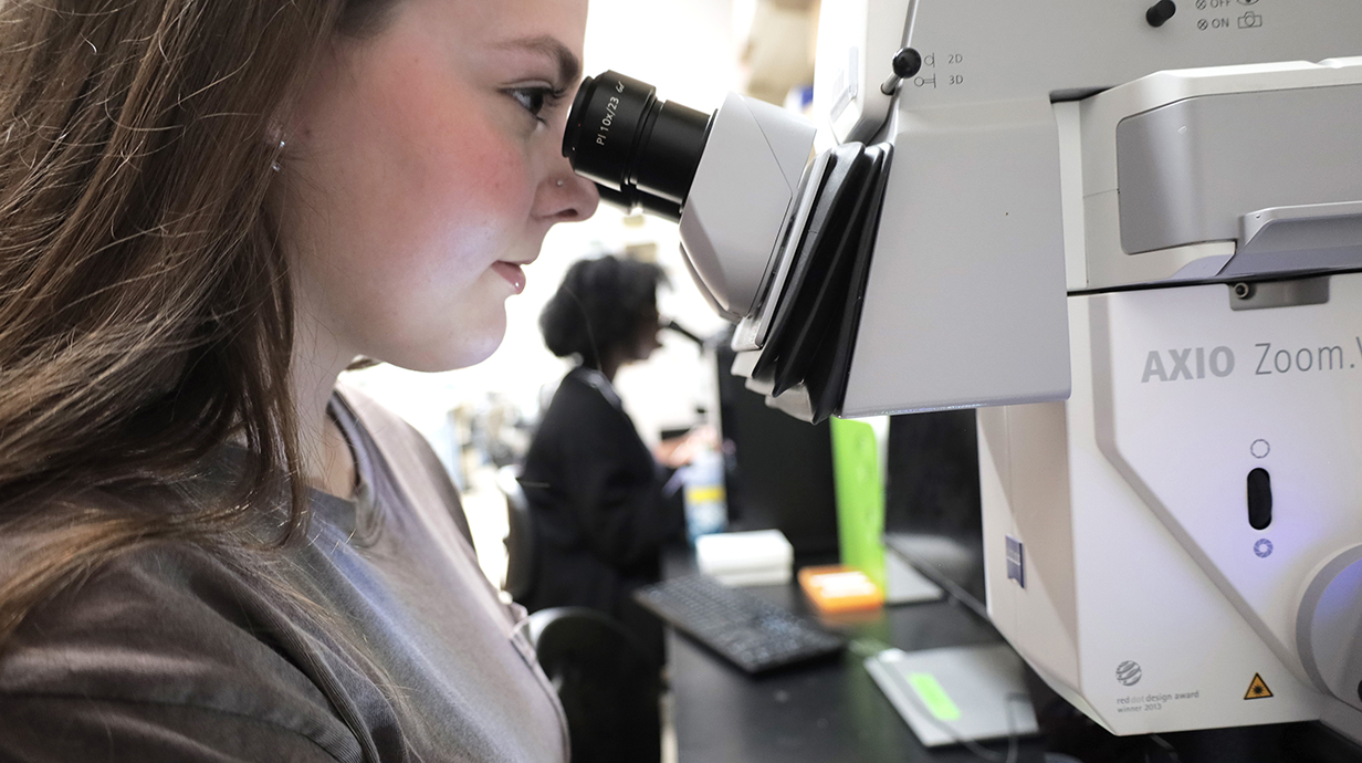 A young woman looks through a microscope in a lab.