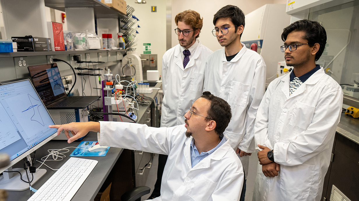 A seated man wearing a white lab coat points to a computer screen in a laboratory while three young men wearing lab coats watch.
