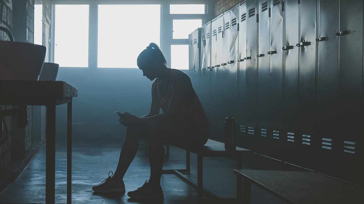 A young woman sits slouched over on a bench in a darkened locker room.