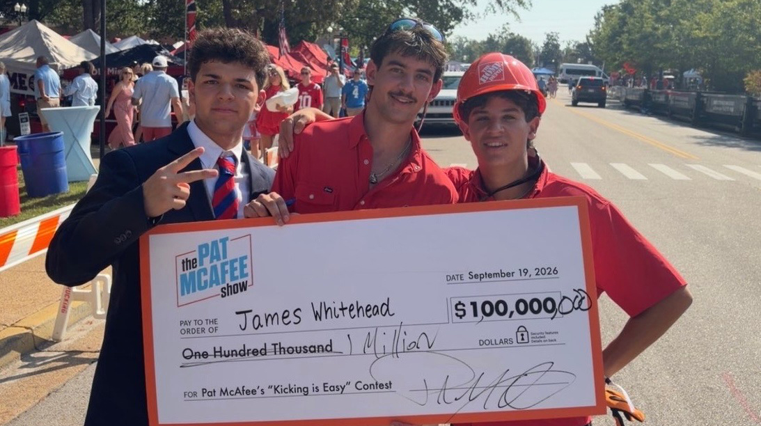 Three young men hold a giant check for $1 million on a street beside a tree-lined park.