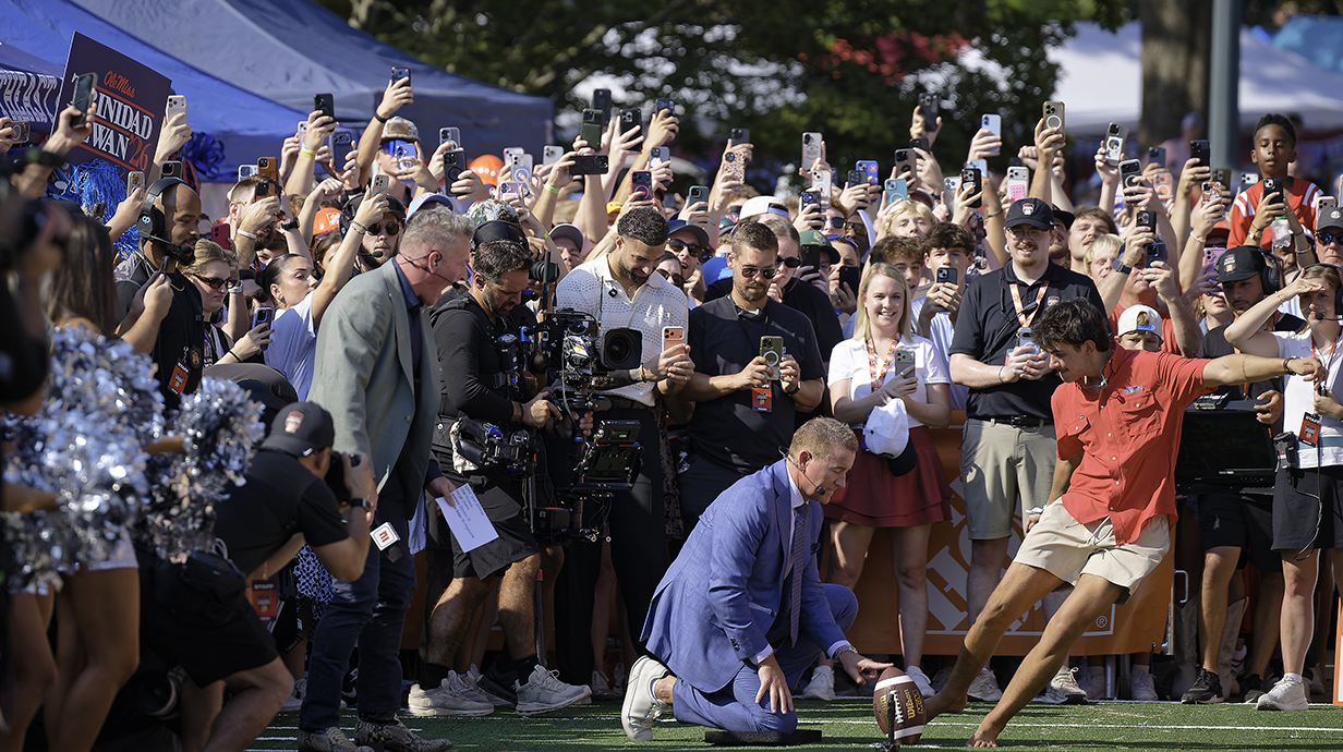 A young man kicks a football barefoot as a large crowd and camera crew cheer him on.