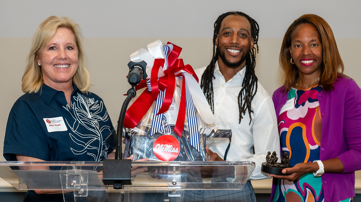 Two women present a wrapped gift and a small trophy to a man with braided hair.