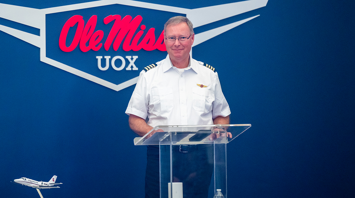 A man wearing a pilot's uniform speaks at a podium in front of a sign reading 'Ole Miss: UOX.'