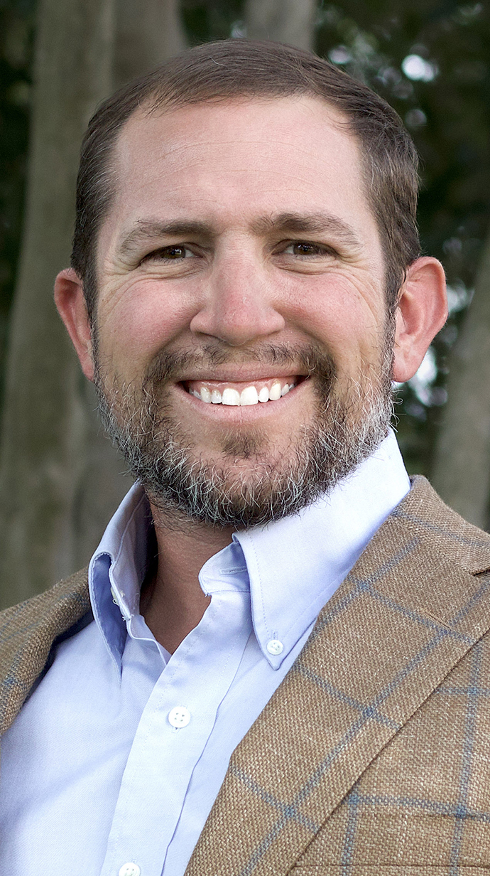 Headshot of a man wearing a tan jacket over a light open-collared dress shirt.