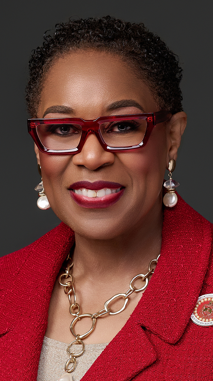 Headshot of a woman wearing a red jacket and gold necklace over a tan blouse.