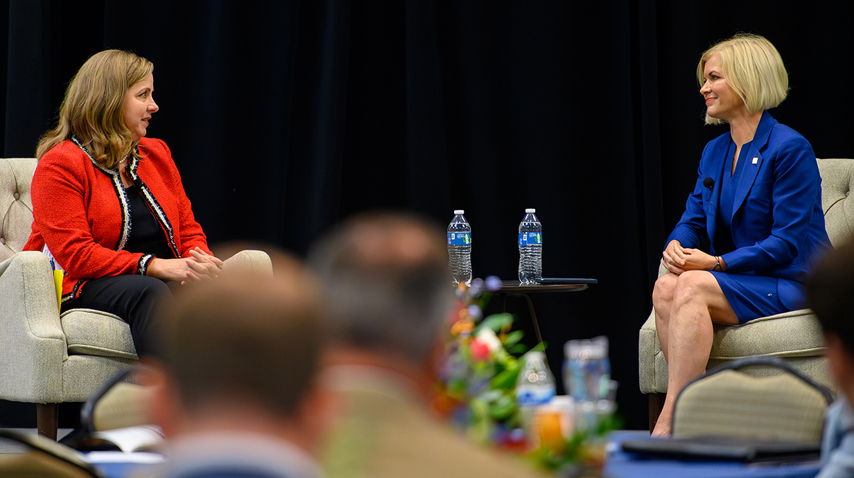 Two women sit in chairs on a stage in front of a crowd in a conference hall.