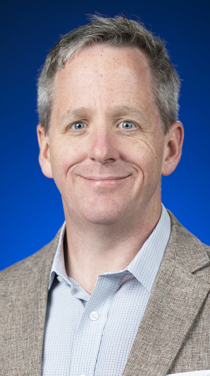 Headshot of a man wearing a tan suit jacket over a light blue shirt.