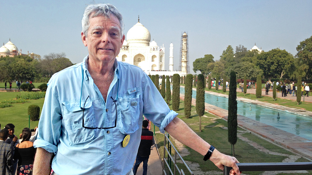 A man wearing a casual blue shirt stands in front of the Taj Mahal.