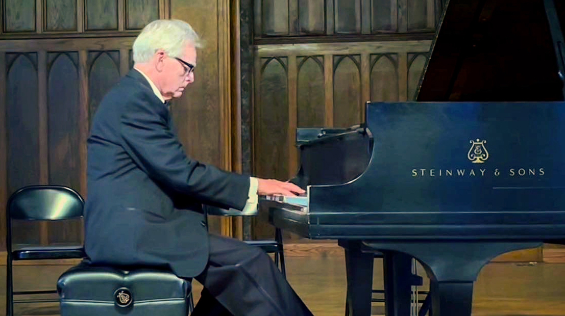 A man wearing a dark suit plays a piano on a stage.