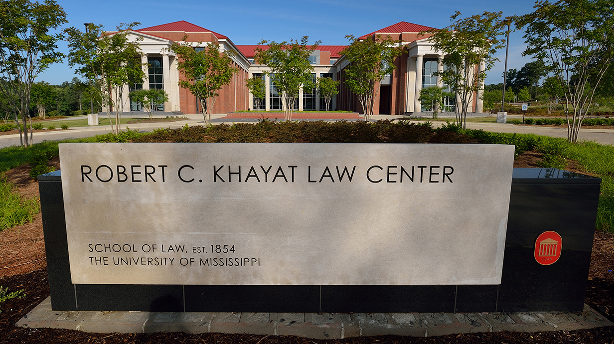 A large stone sign reading 'Robert C. Khayat Law Center' stands in front of a large brick-and-glass building.