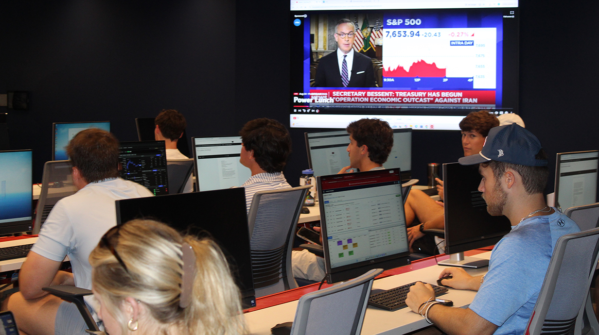 Several young men and women sit at computer terminals in a darkened room with a television displaying financial news on the back wall.