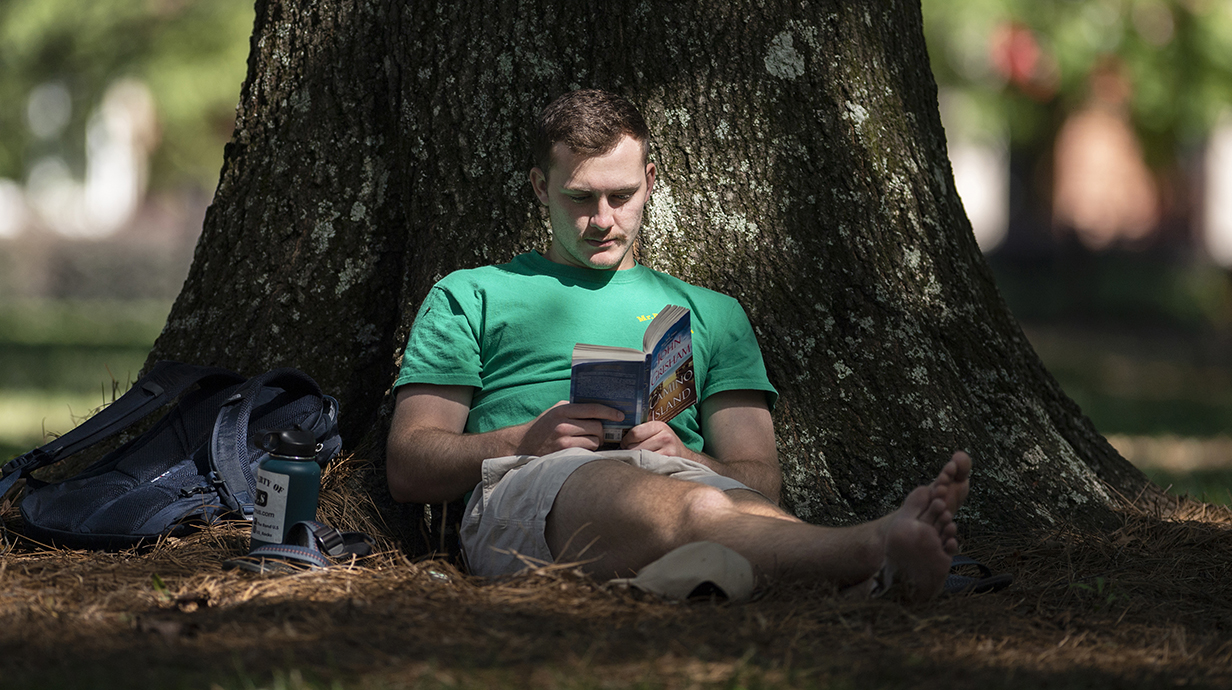 A young man reads a book while sitting in the shade at the base of a large tree.