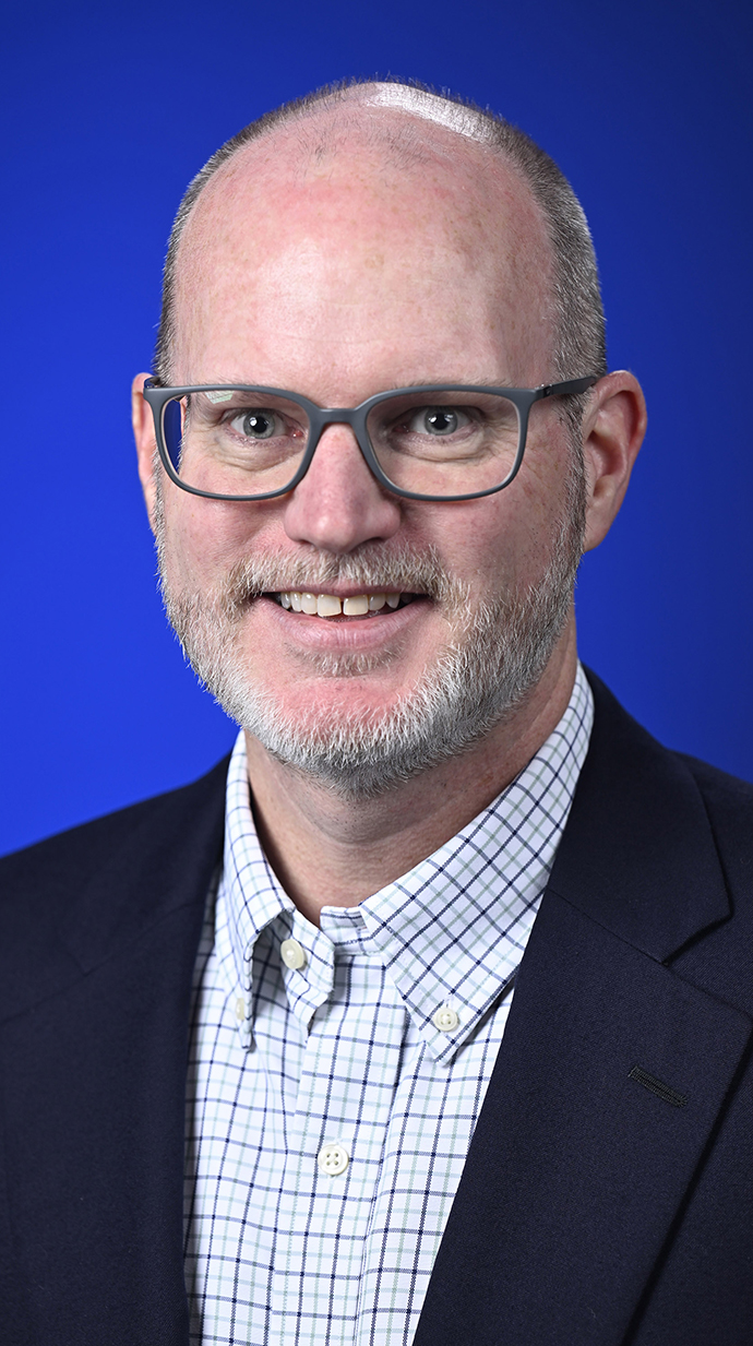 Headshow of a man wearing a dark bluw jacket over a shirt with a blue windowpane checked pattern.