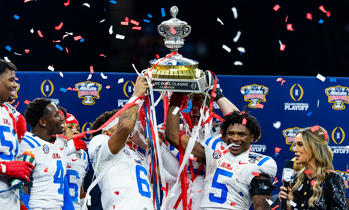 Football players celebrate with a large trophy in a domed arena.