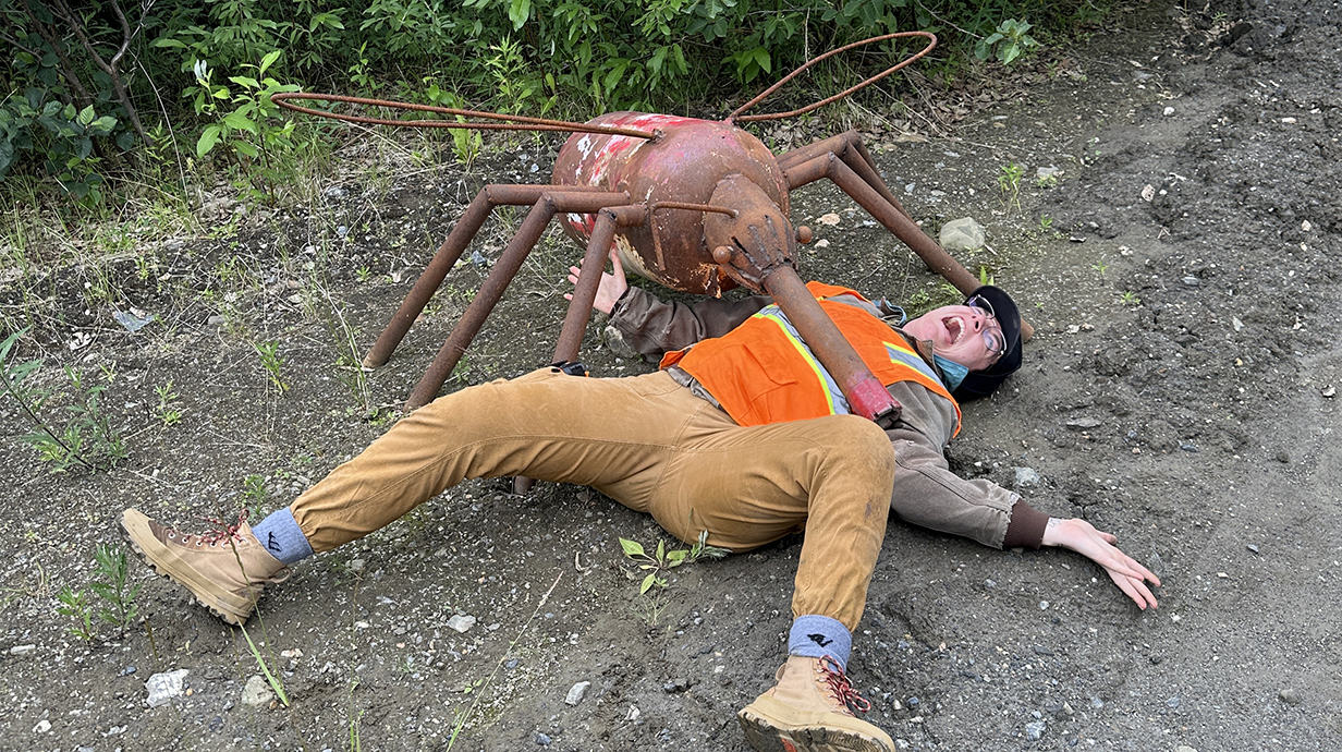 A woman wearing an orange safety vest lies on the ground beside a large metal sculpture of a mosquito.