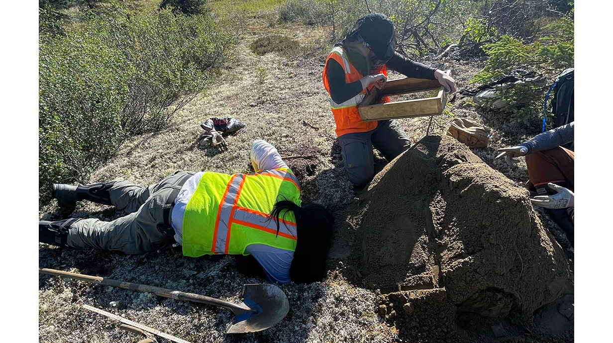 A young woman wearing a mosquito net and orange safety vest sifts dirt while a man wearing a yellow vest lies on the ground and looks into a hole.