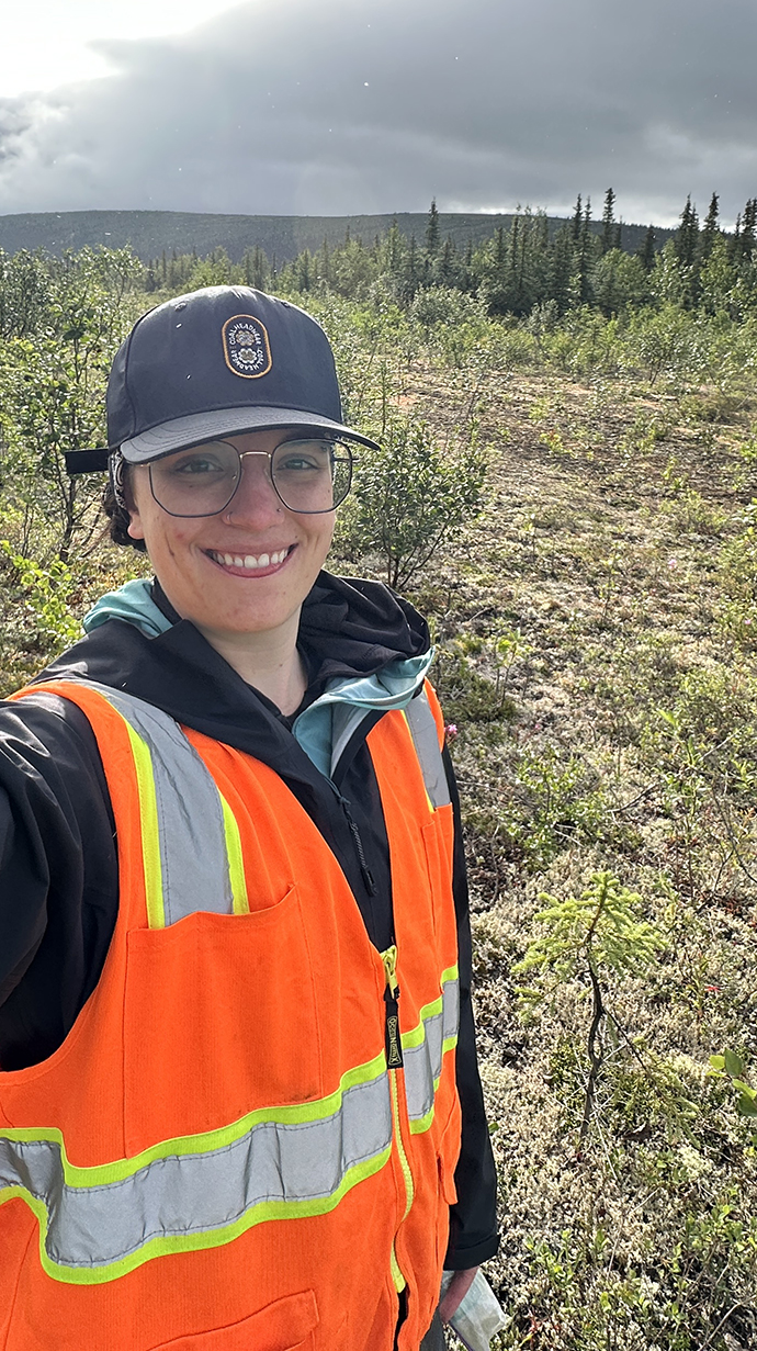 A young woman wearing an orange safety vest stands in a mountain valley with little vegetation.