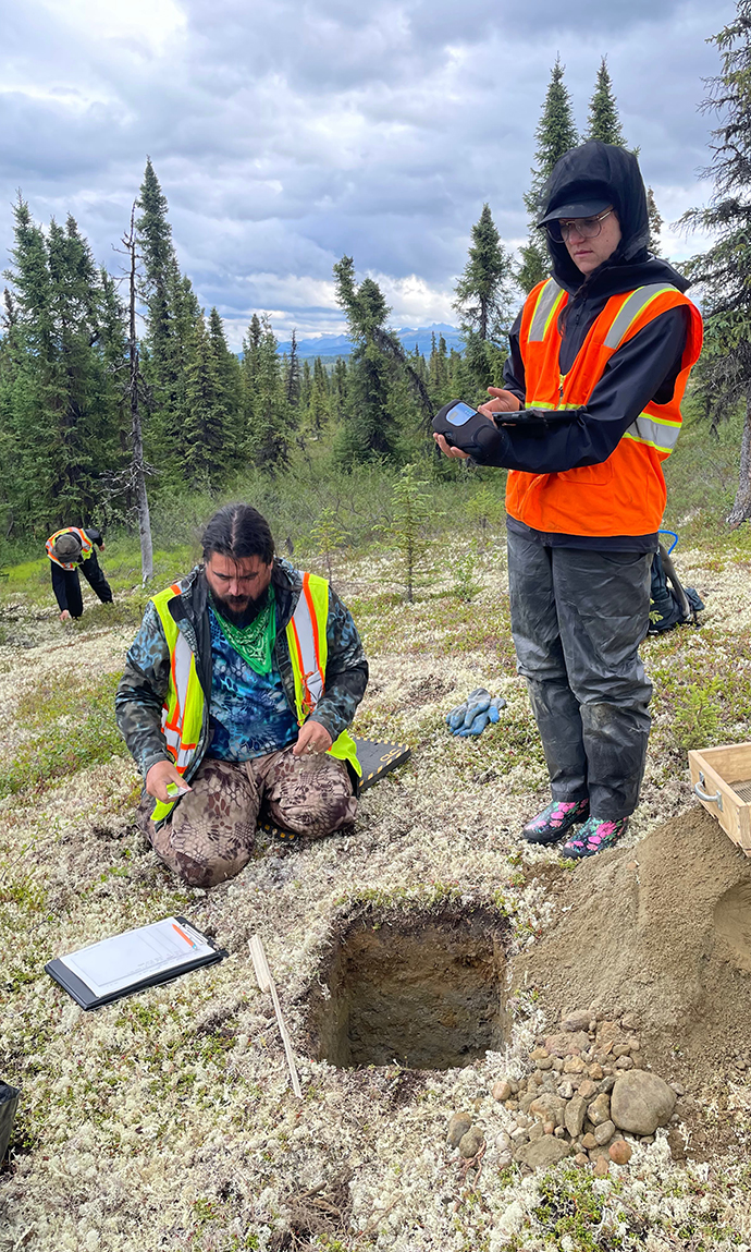 A young woman wearing an orange safety vest types into a tablet computer at an outdoor site as a man looks into a hold in the bare groung.