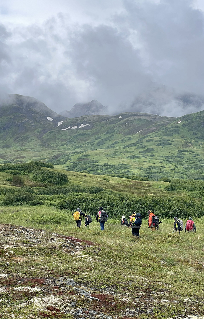 A group of people hikes through a valley in front of cloud-topped mountains.