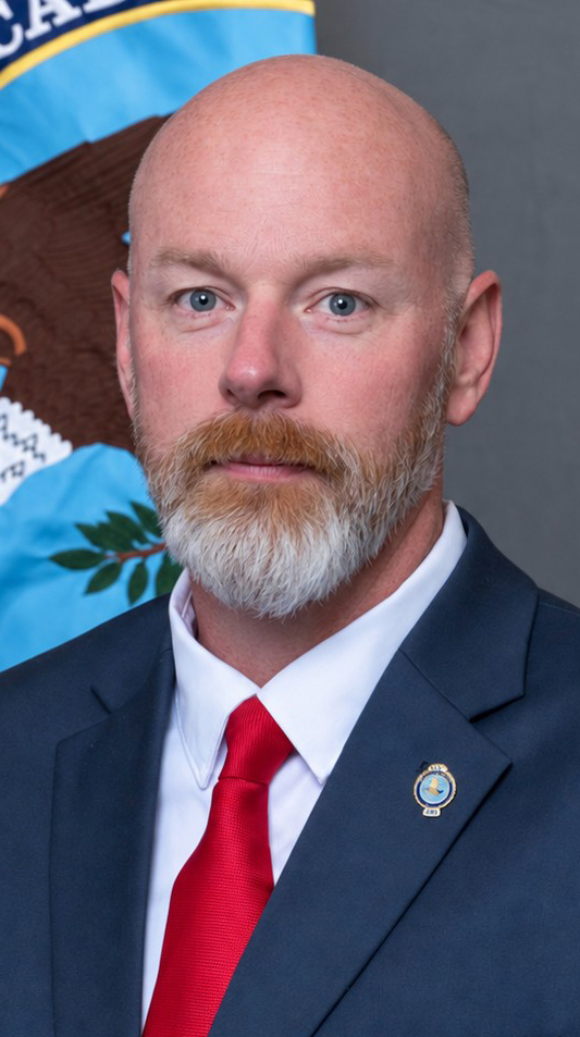 Headshot of a man wearing a blue suit and a red tie standing in front of a colorful flag.