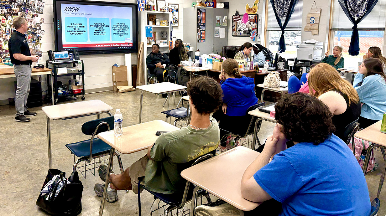 A man standing beside a projection screen talks to a classroom full of students.