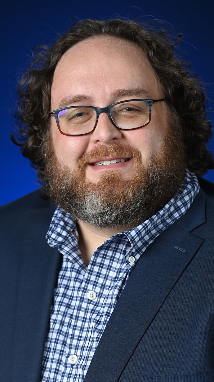 Headshot of a man wearing a dark blue jacket over a blue patterned dress shirt.