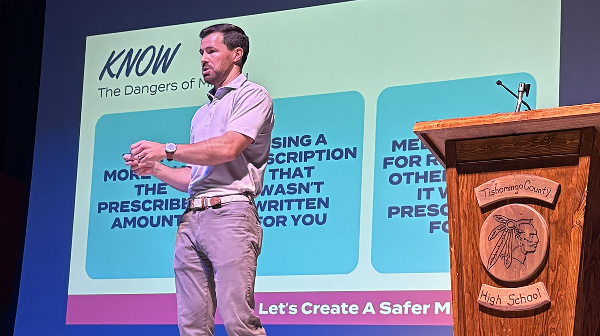 A man speaks while standing in front of a projection screen.