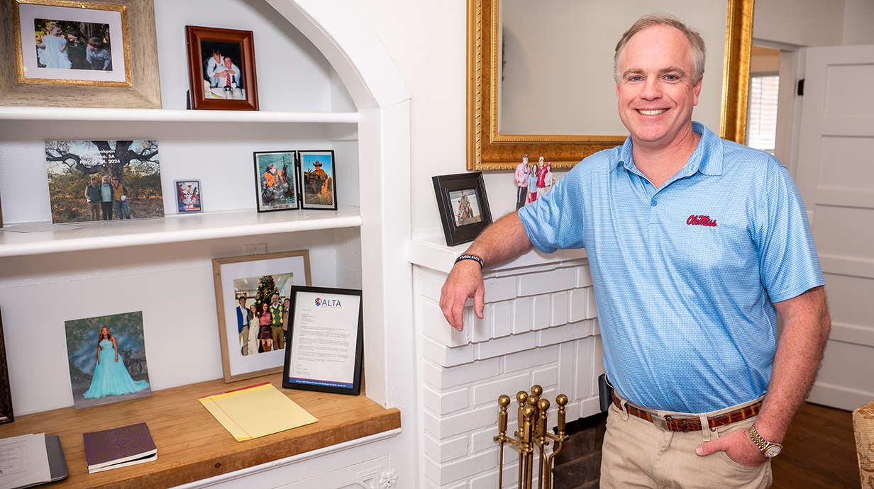 A man wearing a blue Ole Miss polo shirt leans against built-in shelves filled with family photos.