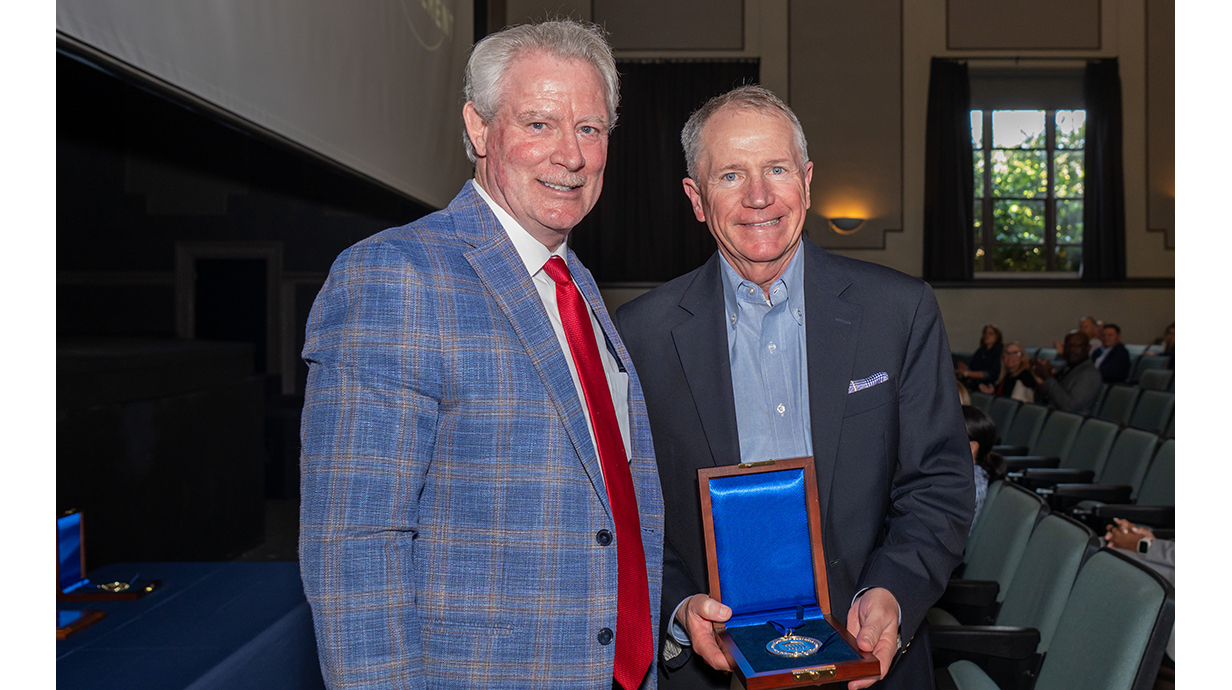 A man wearing a blue suit presents another man with a medal in an auditorium.