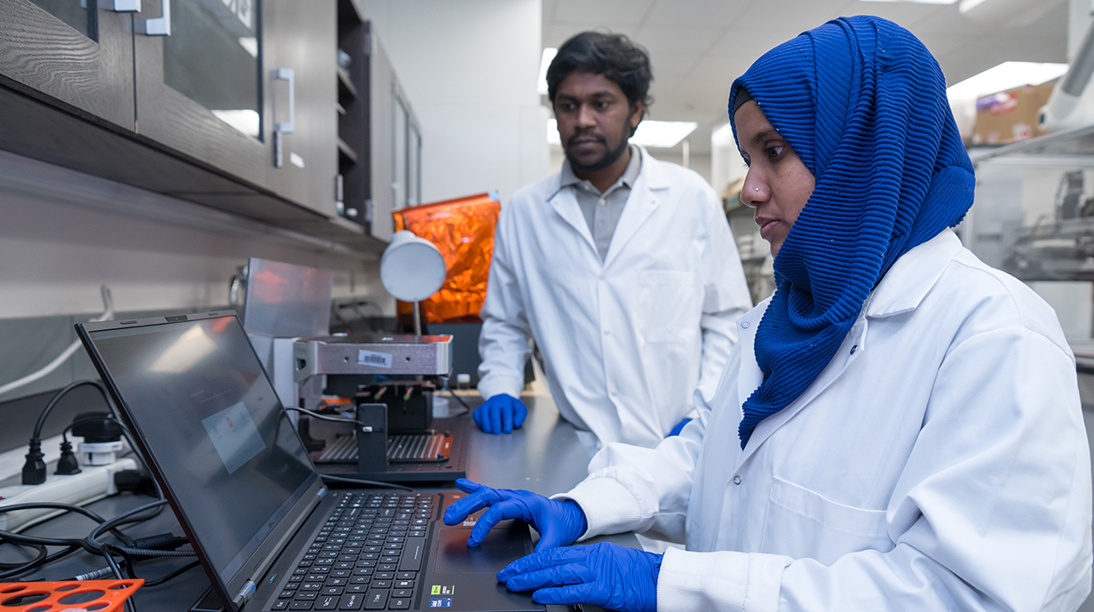 A young man wearing a white lab coat and a young woman wearing a white lab coat and a blue head scarf work with equipment in a lab.