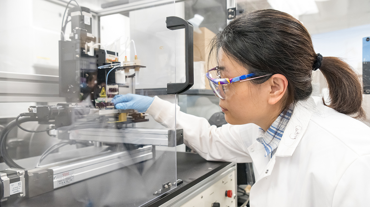 A young woman wearing a white lab coat and satery glasses adjusts a 3D printer in a lab.