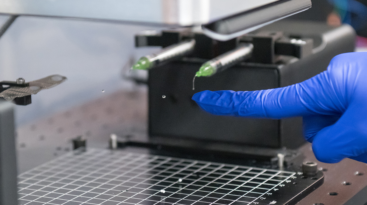 A hand wearing a blue latex glove points at a tiny droplet of liquid suspended in midair in a lab.