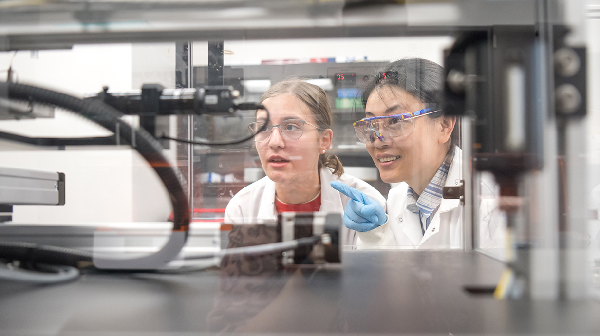 Two women wearing white lab coats look inside a 3D printer as it works.