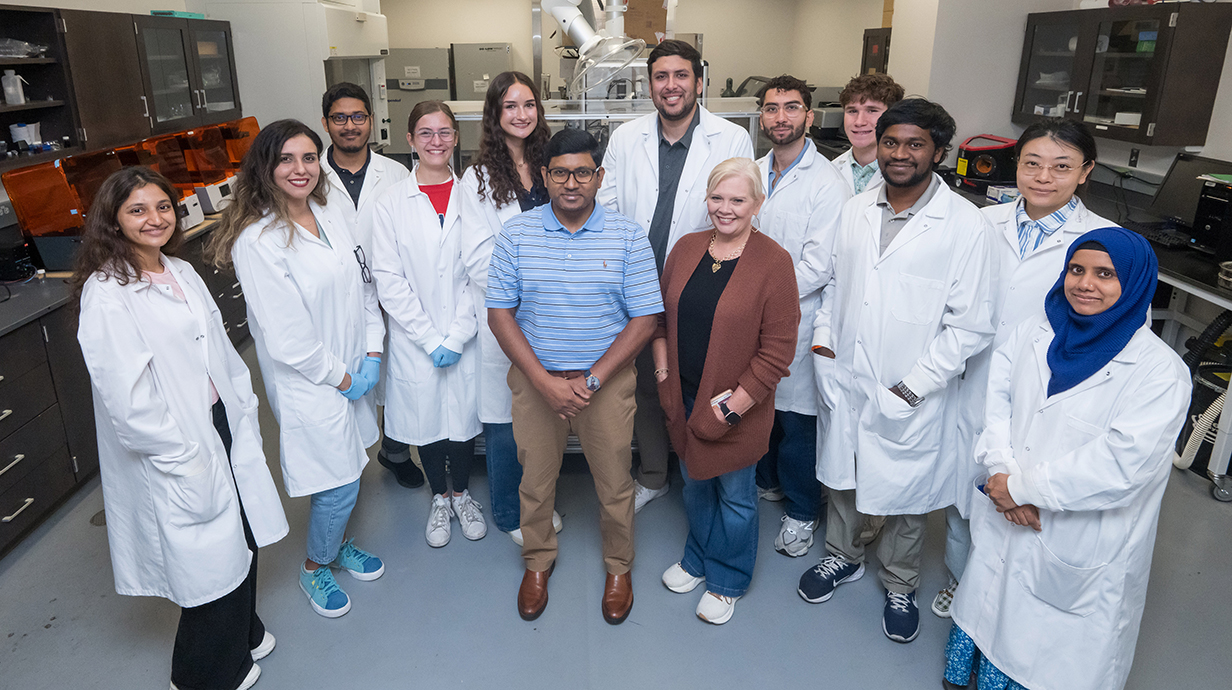 A large group of young men and women, most wearing lab coats, stand in front of equipment in a pharmaceutical laboratory.