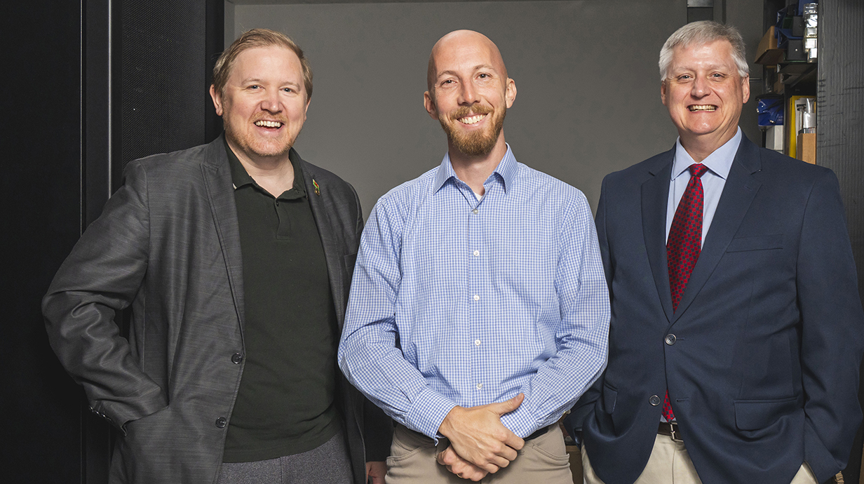 Three men stand in a room lined with racks of computer servers.