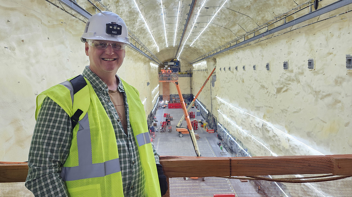 A man wearing a hardhat and safety vest stands in a cavern filled with construction equipment.