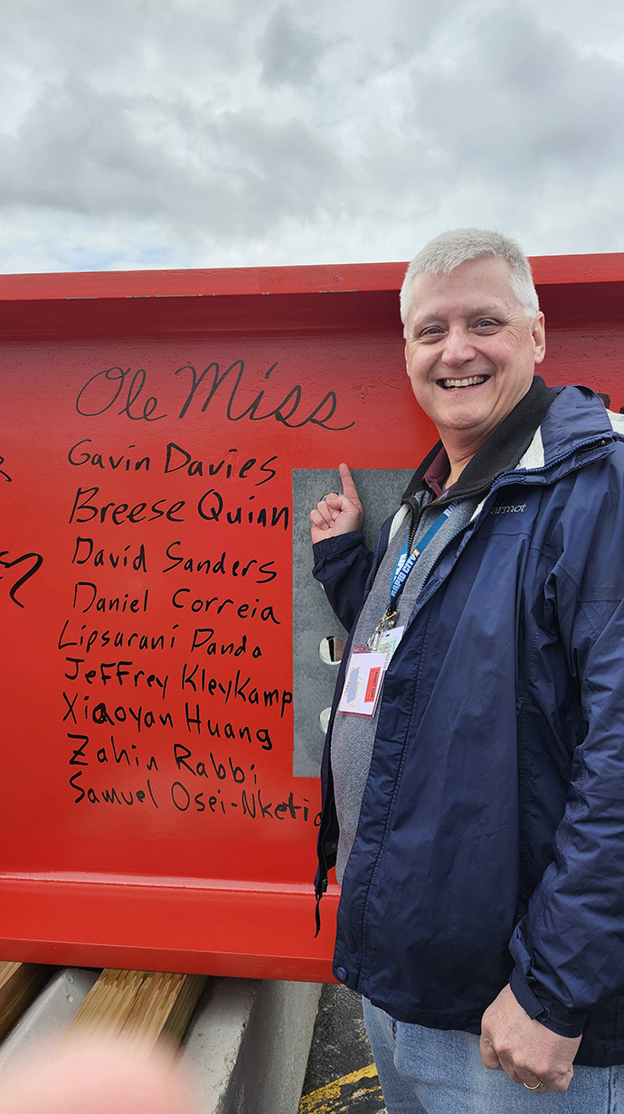 A man points to several signatures on a bright red steel beam.