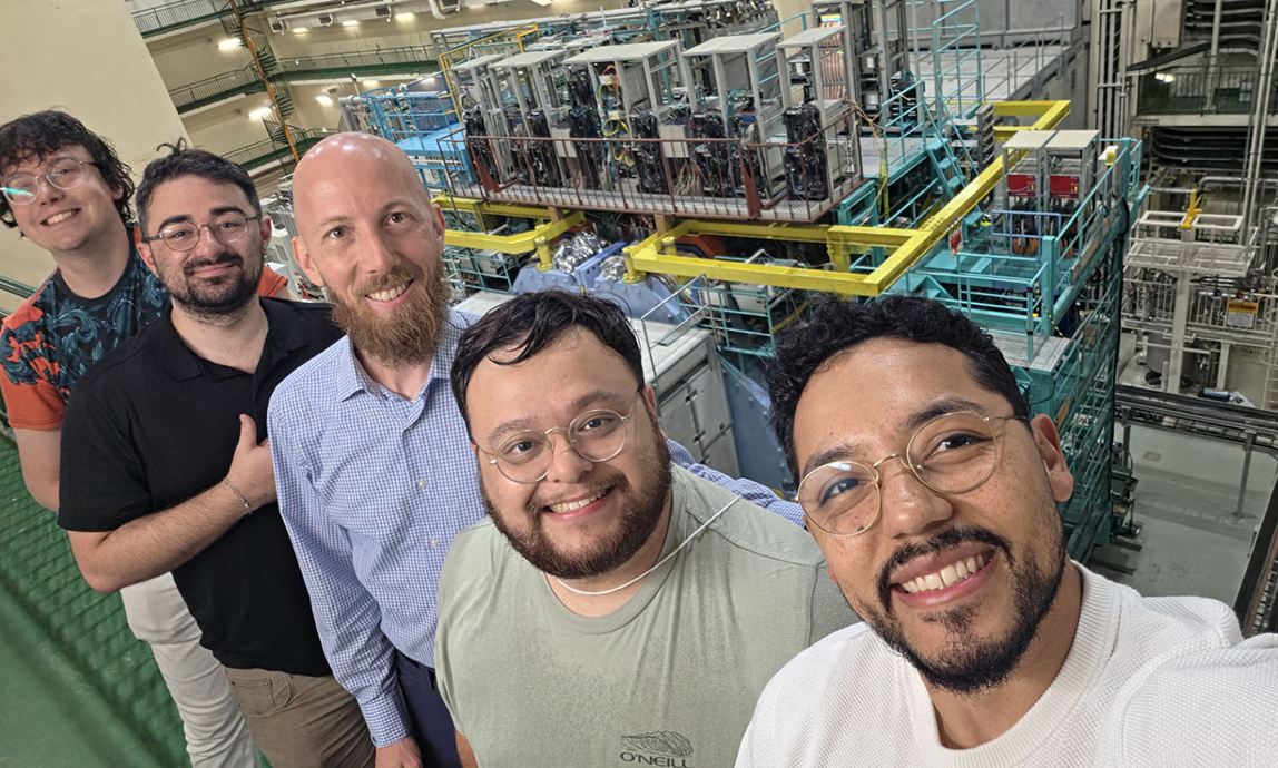 Five men pose for a photo while standing on a catwalk above a giant scientific instrument.