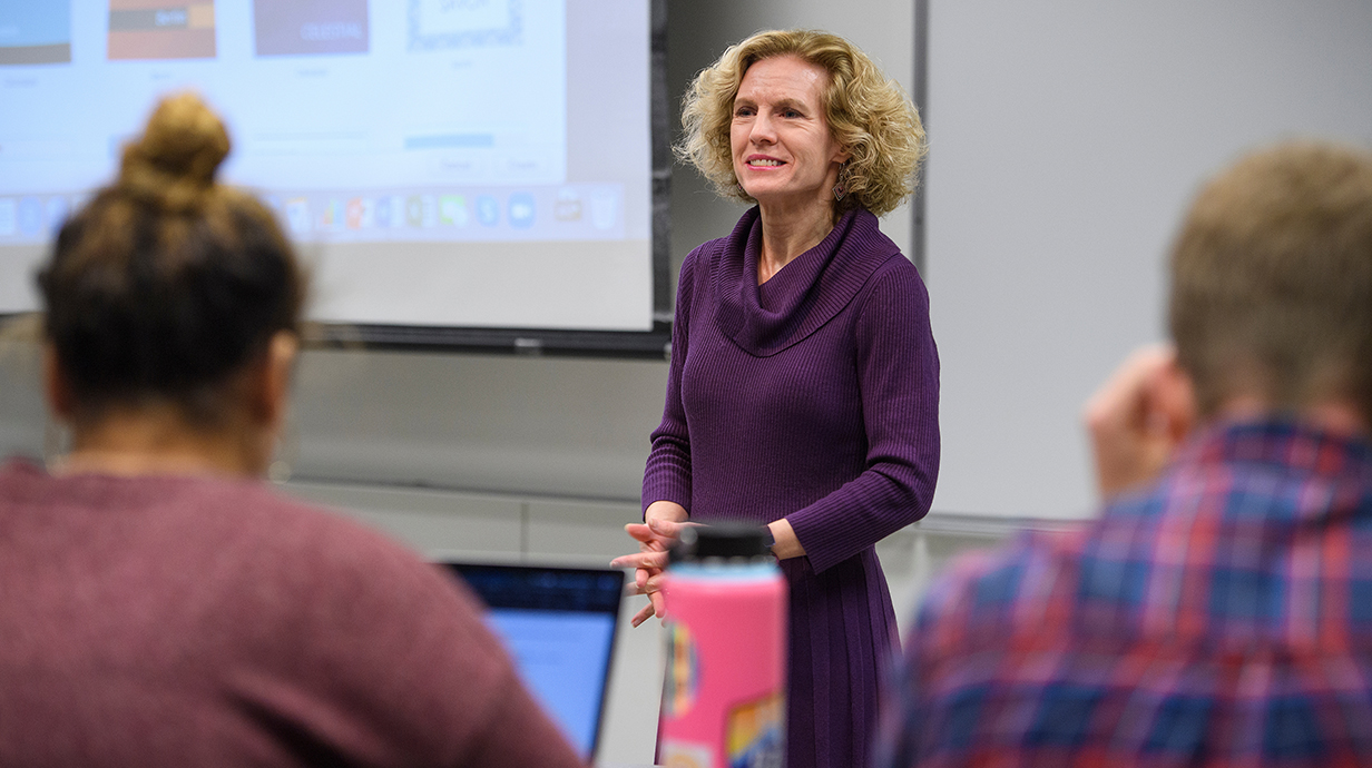 A woman wearing a purple dress speaks to students in a college classroom.