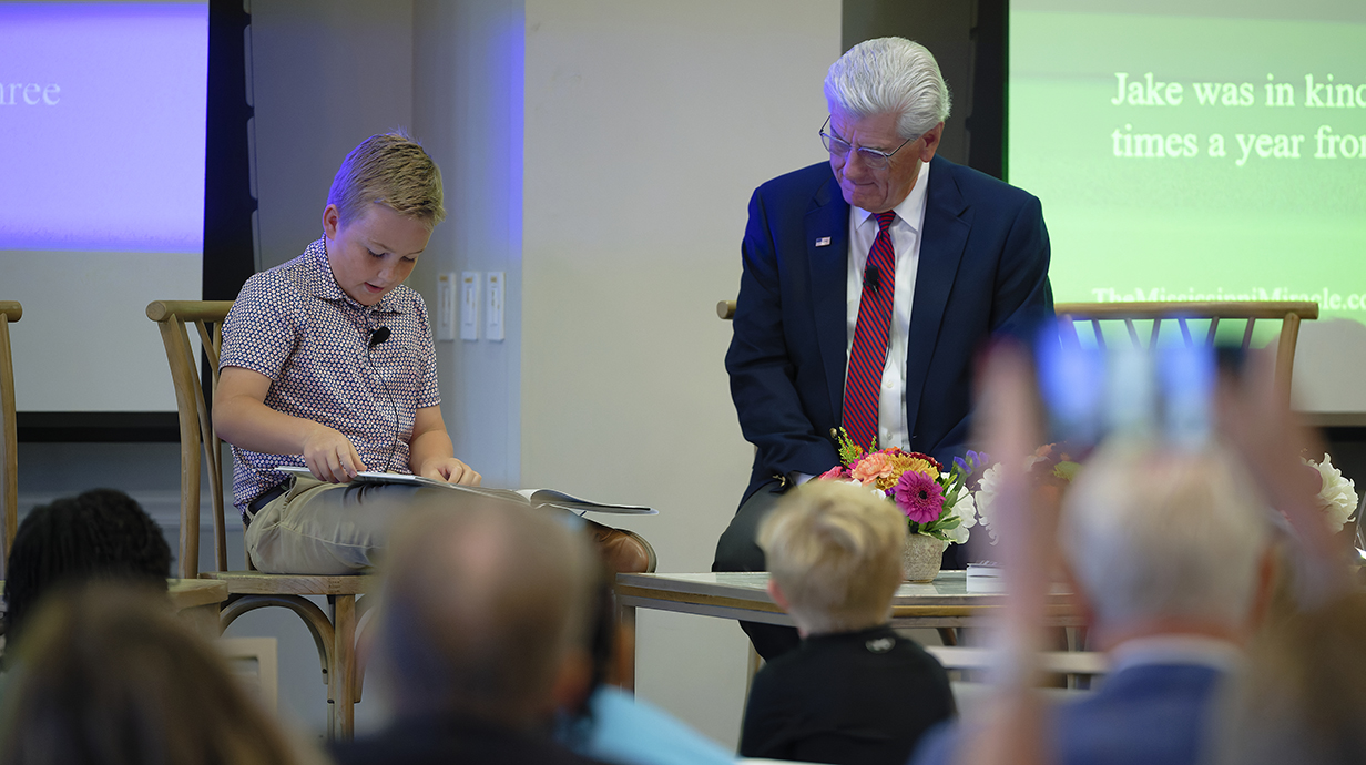 A young boy reads a book while sitting on a stage as a man seated nearby looks on.