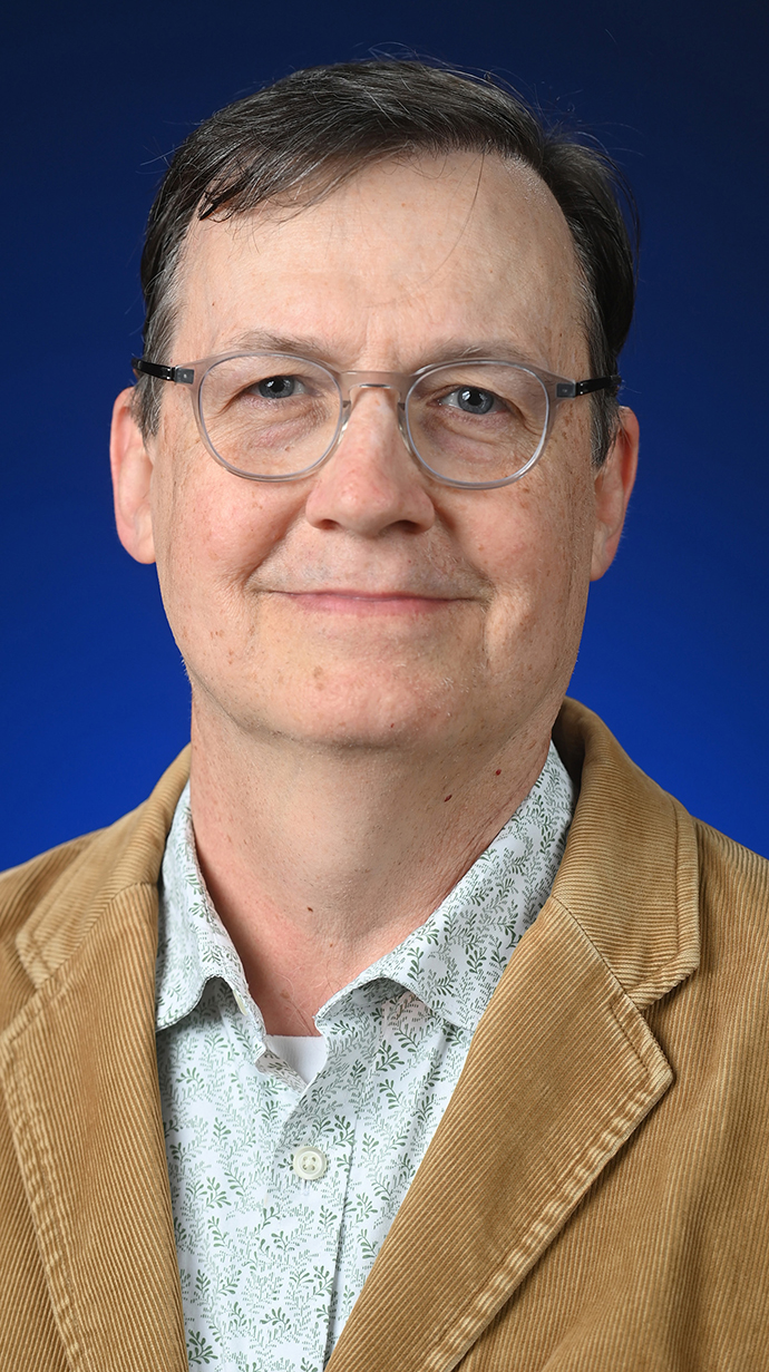 Headshot of a man wearing a tan jacket and a green-and-white patterned dress shirt.