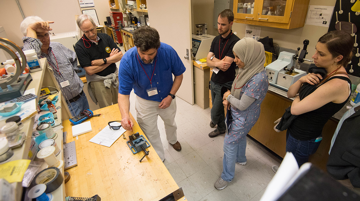 A man demonstrates a piece of equipment in a vise for a small group of people visiting a laboratory.