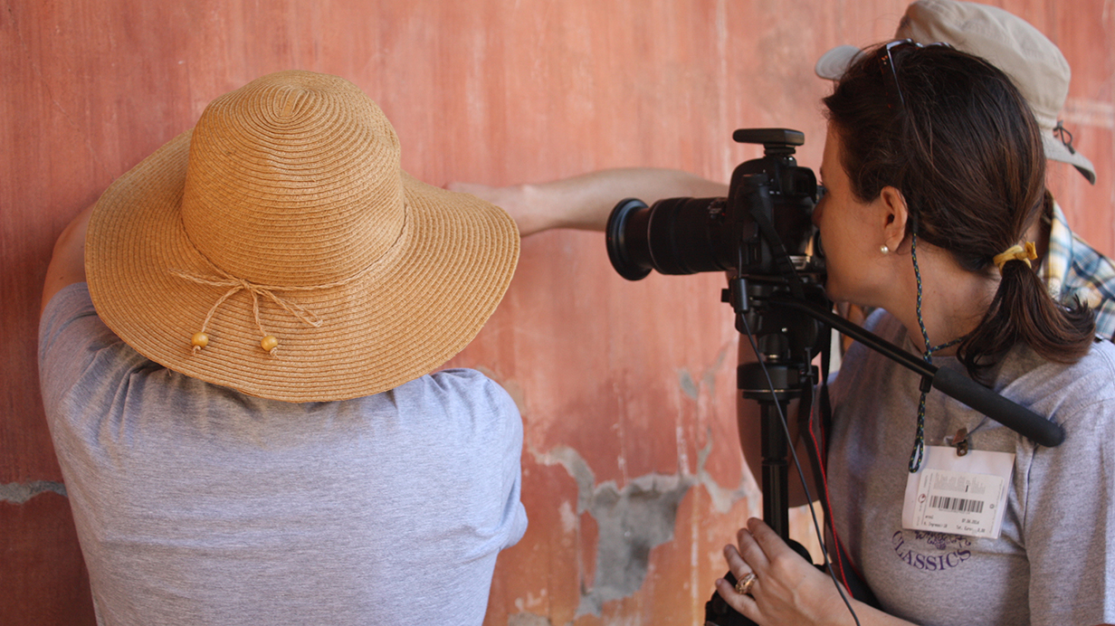 A woman uses a digital camera to photograph a section of a pink wall that another women points at.
