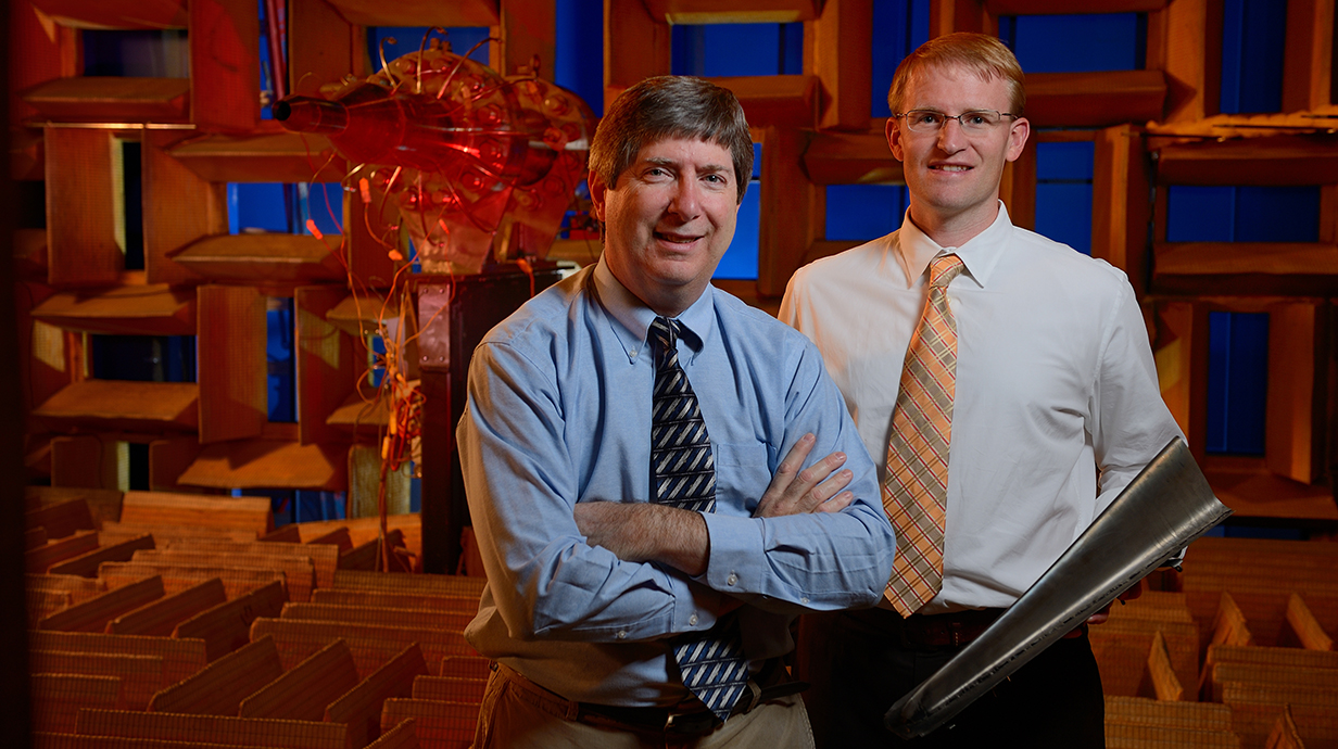 Two men hold a piece of metal in front of a jet engine mock-up in an acoustics laboratory.