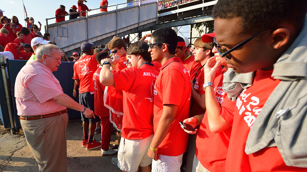 A man wearing a pink shirt talks to a group of young people, all wearing red T-shirts.