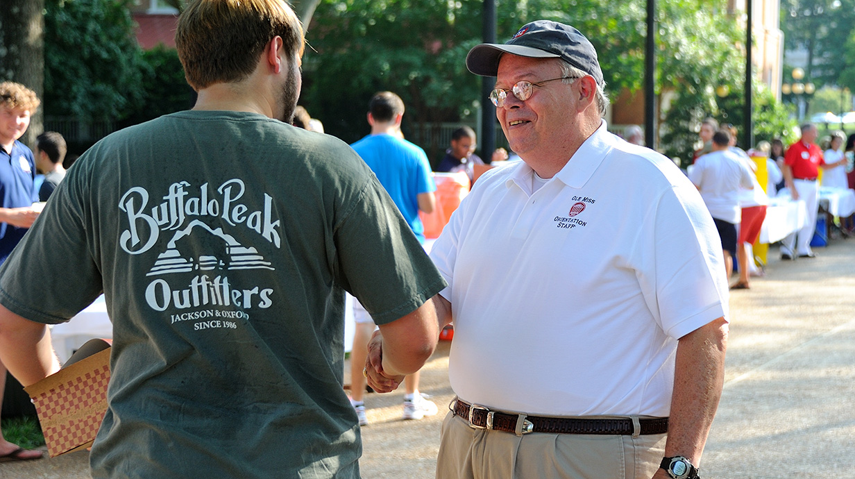 A man wearing a white polo shirt talks to a young man wearing a green T-shirt at an outdoor event.