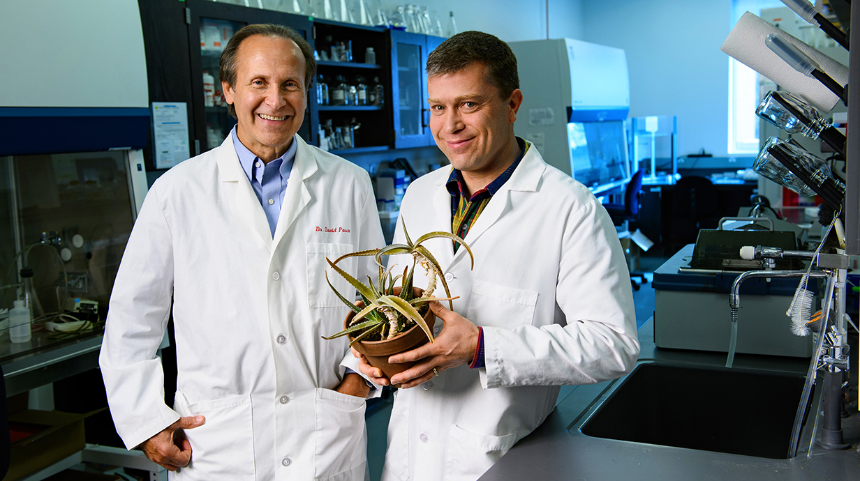 Two men wearing white lab coats, one holding a potted aloe vera plant, stand in a laboratory.