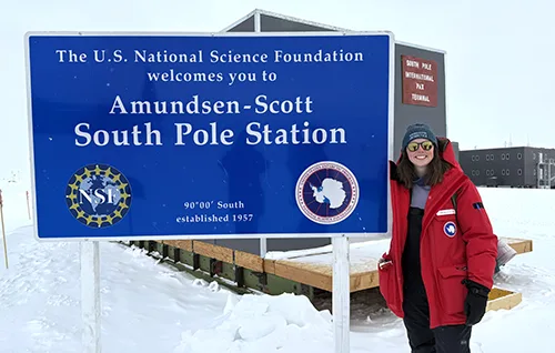 person standing in bright red coat and hat next to sign that reads:The U.S. National Science Foundations welcomes you to Amundsen-Scott South Pole Station. Snowy conditions