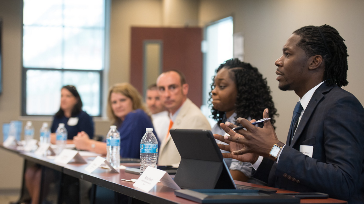 Six educators in professional attire sit on a panel and direct their focus to a man on the end.
