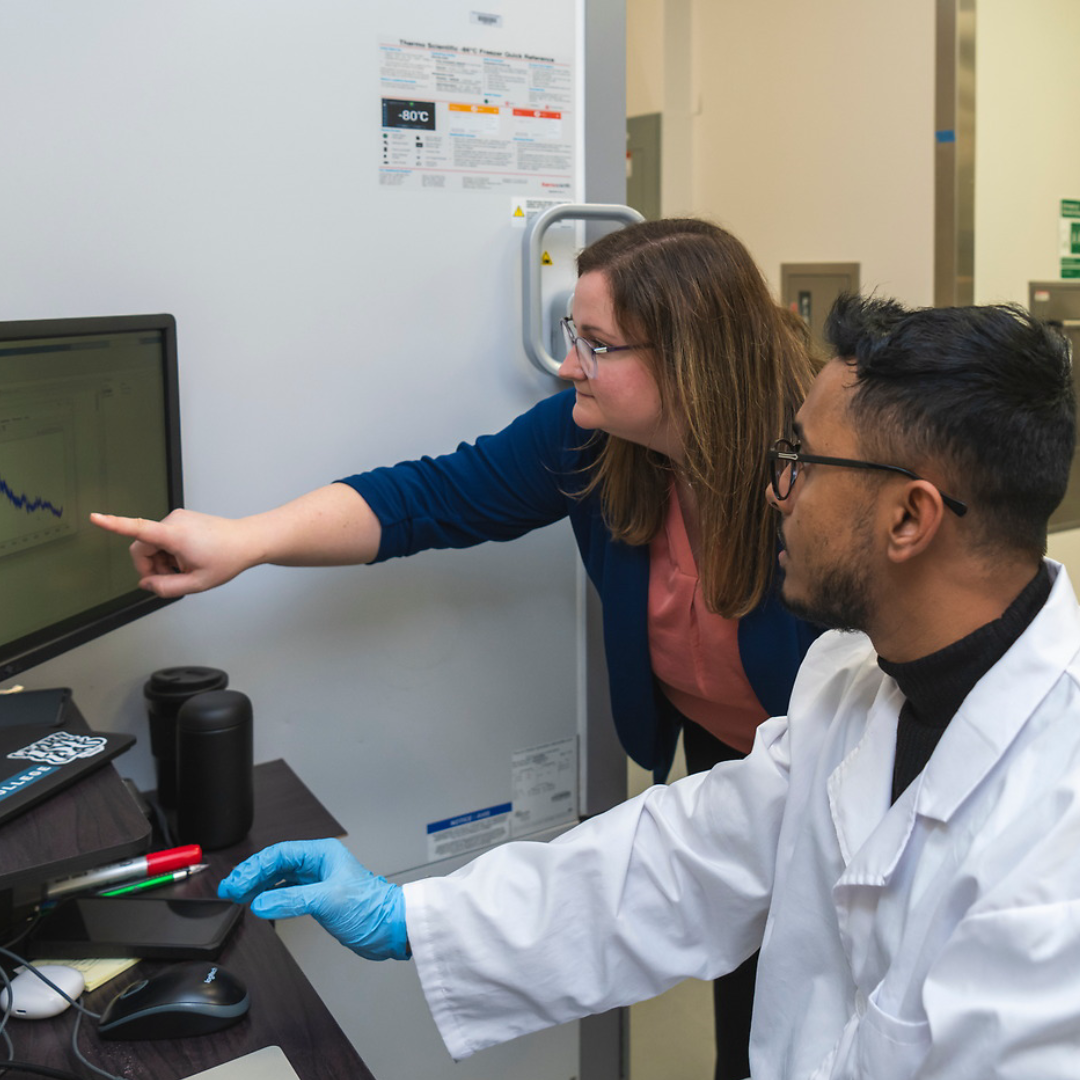 An instructor points to a graph on a computer while a researcher in a lab coat sits at the computer looking intently